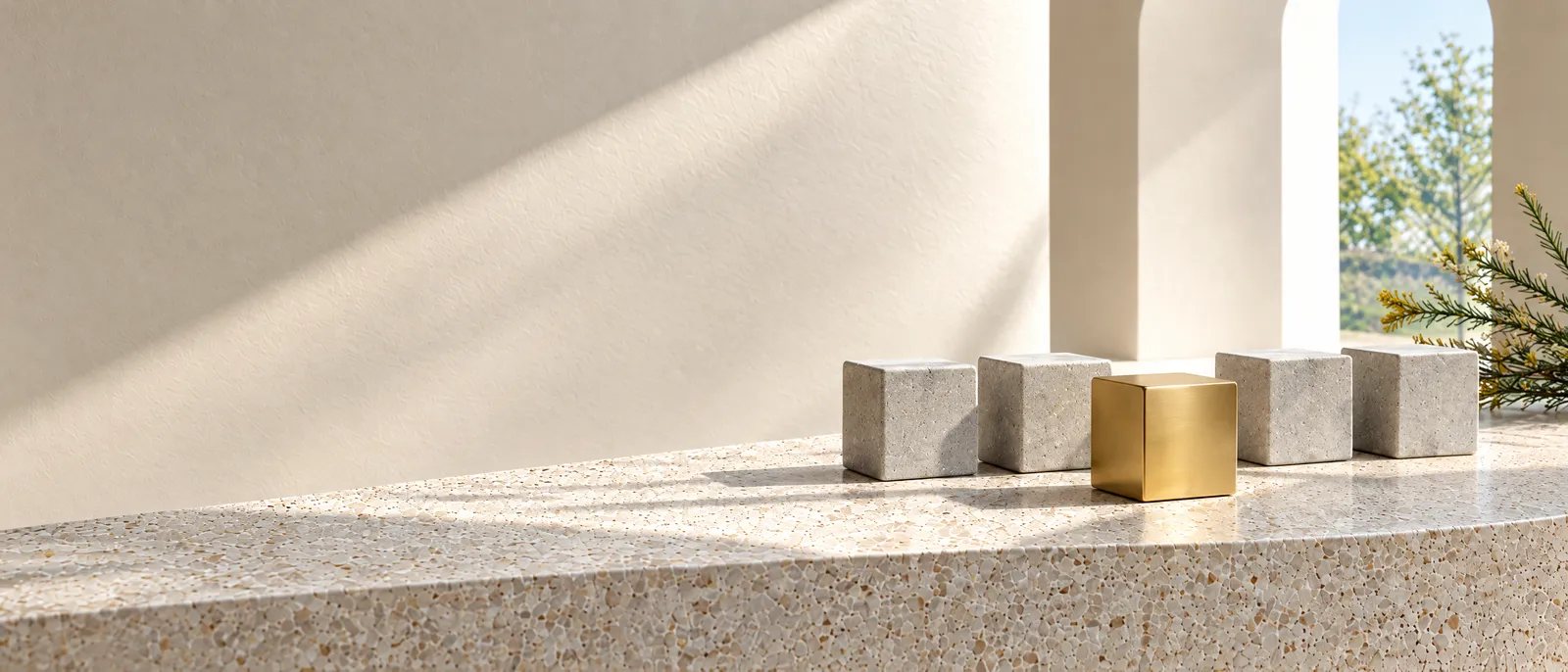 A row of plain grey stone cubes on a pale terrazzo ledge in daylight, one cube cased in brass standing out
