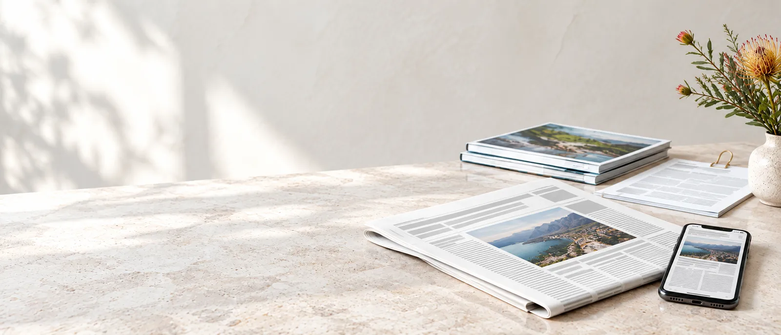 A newspaper open at a feature article on a pale stone table, the same story on a phone beside it, magazines behind