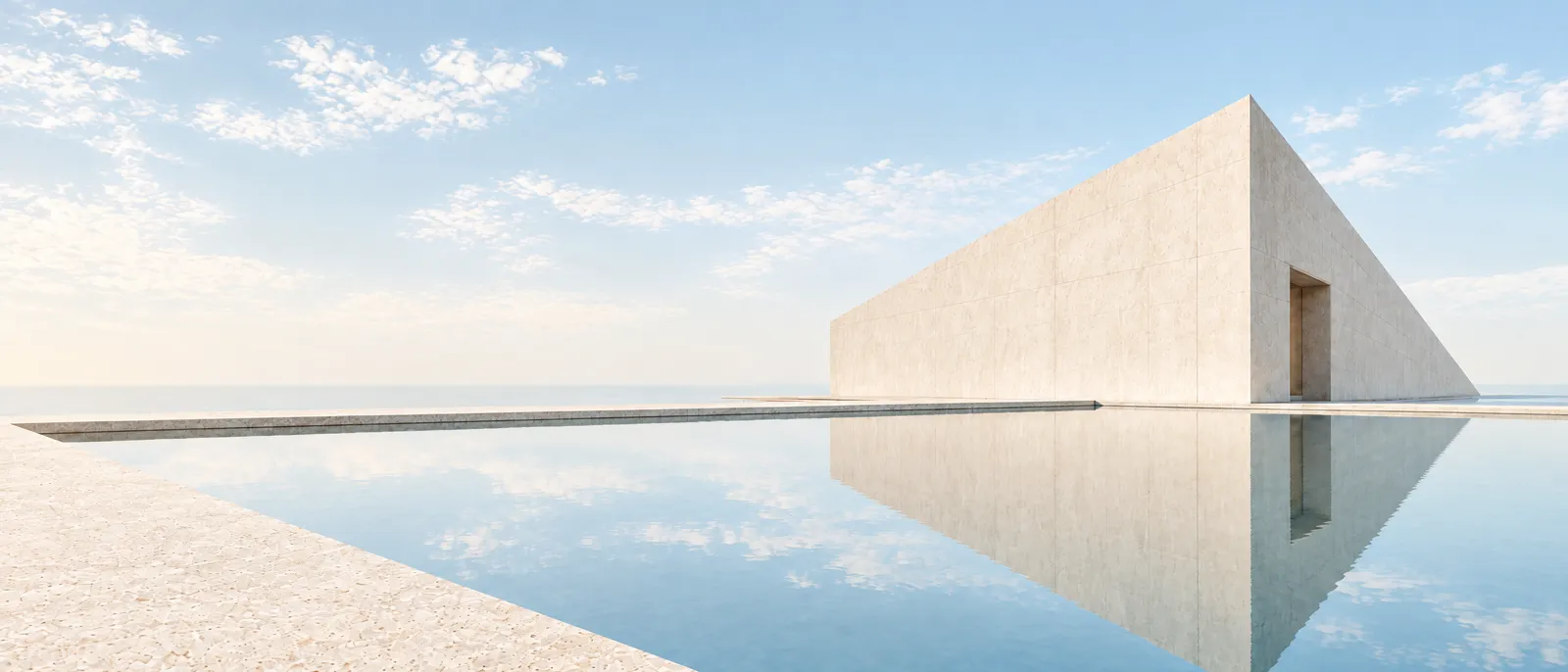 A pale concrete building beside a still pool reflecting a light sky, in soft daylight